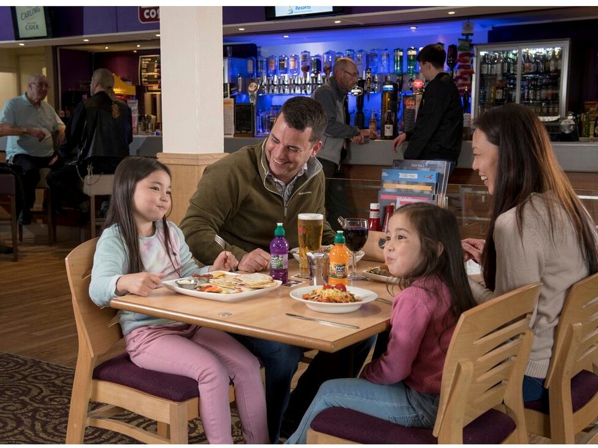 A family of four eating plated meals at a wooden table in a carpeted bar and restaurant, with the lit bar counter behind them.