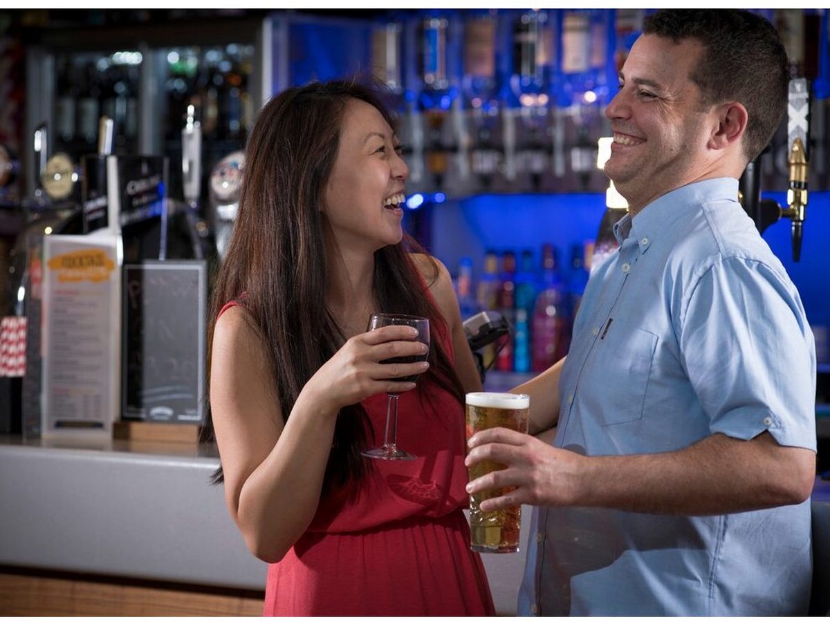 A couple standing at the bar counter with drinks, in front of draught beer taps and backlit bottle shelves.