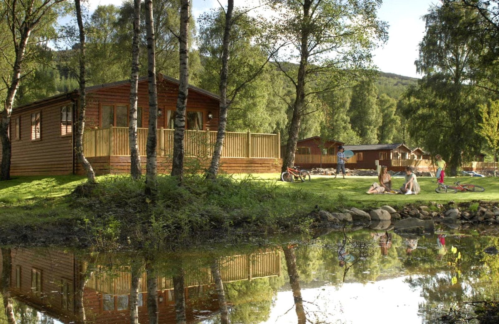 A timber lodge with a decked veranda among birch trees beside a pond at Tummel Valley Holiday Park, with families sitting out on the grass and wooded hills behind.
