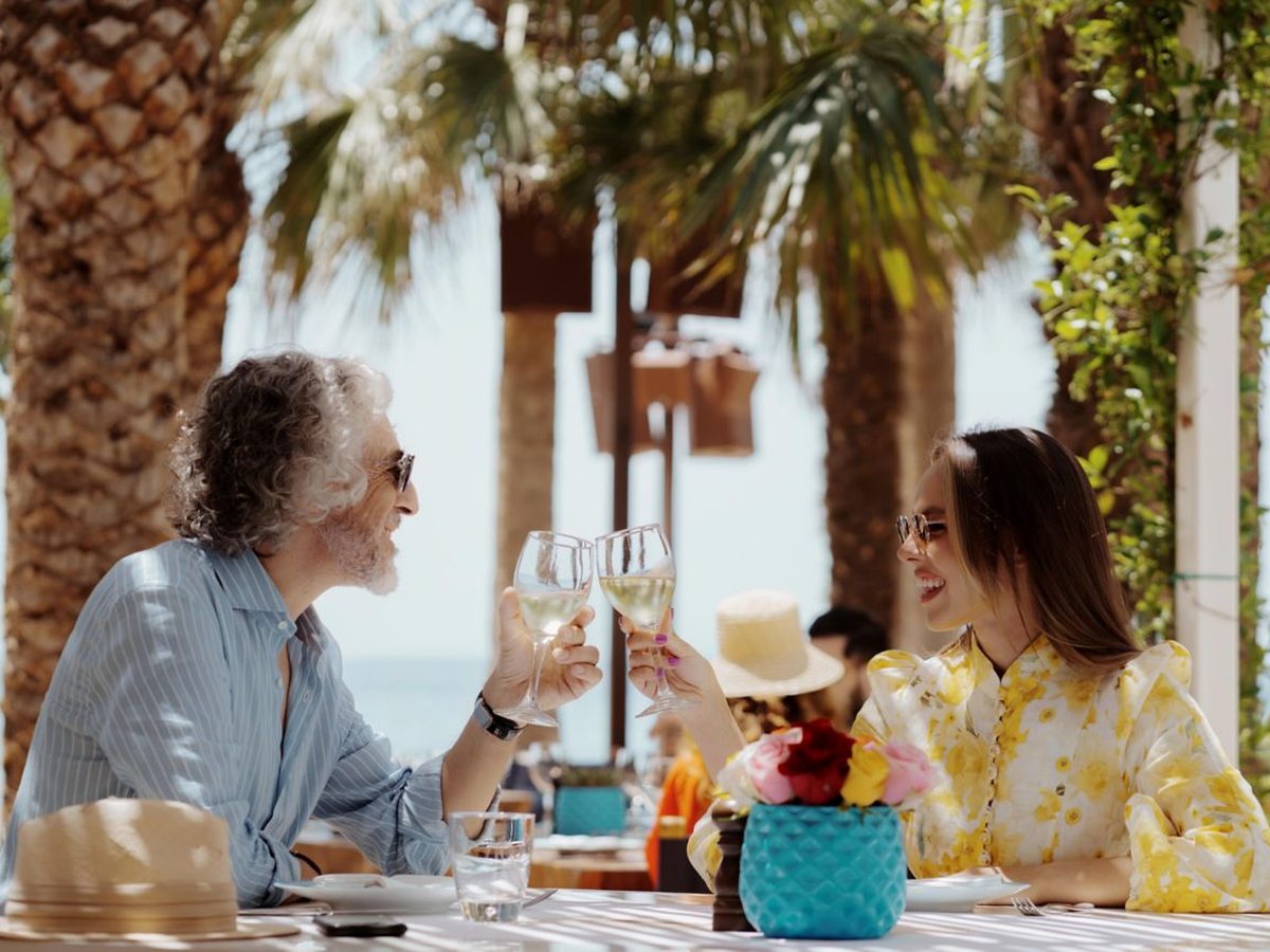 Two guests touching wine glasses across a table on an open-air restaurant terrace, under palm trees with the sea behind them.