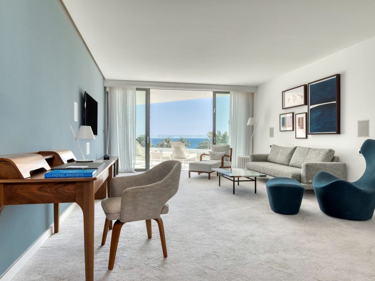 An open-plan suite living area with a grey sofa, blue armchairs, a writing desk and sliding doors onto a furnished sea-view terrace.