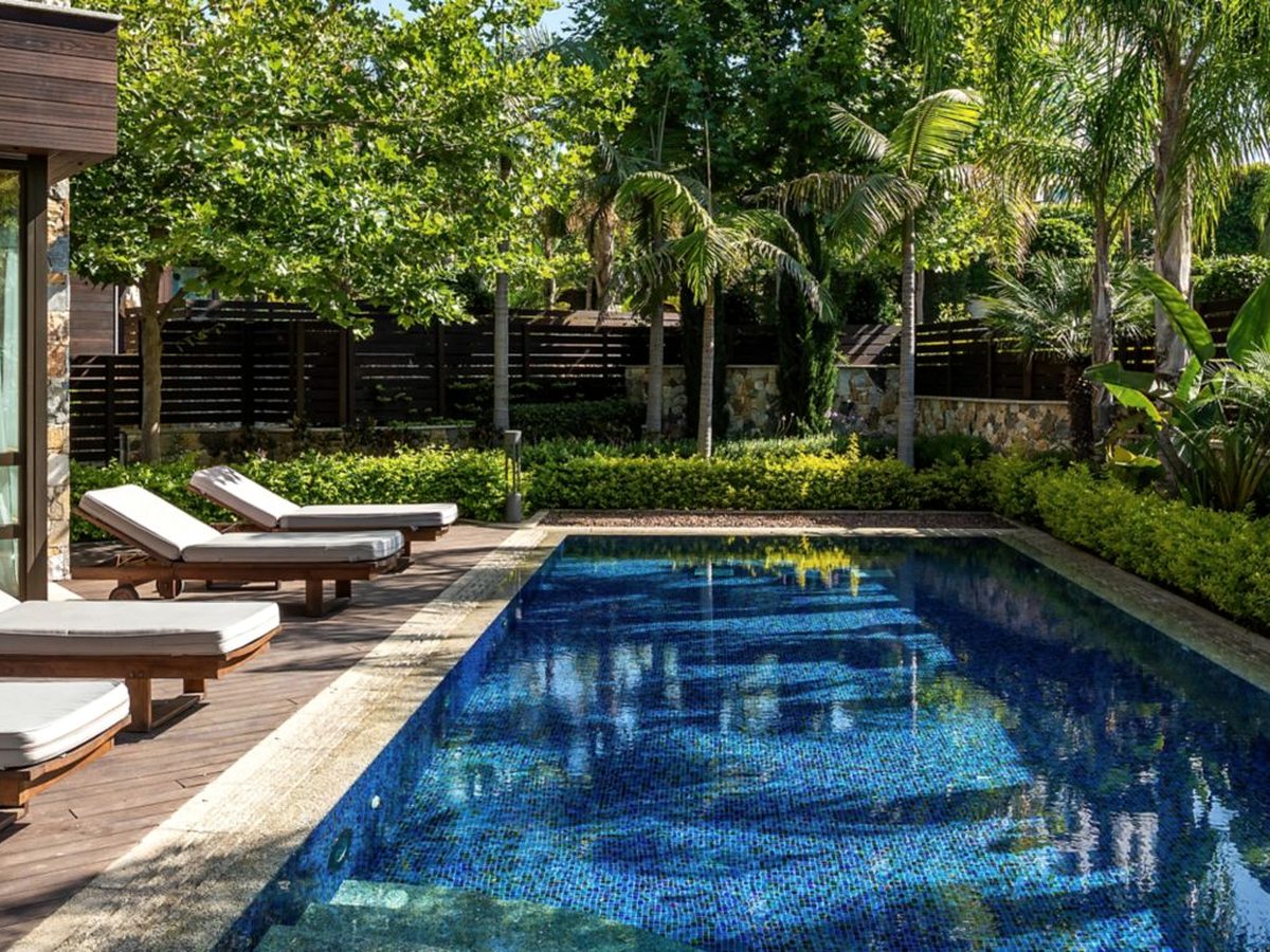 A rectangular mosaic-tiled pool beside a timber deck with wooden sun loungers, enclosed by clipped hedges, palms and a slatted fence.