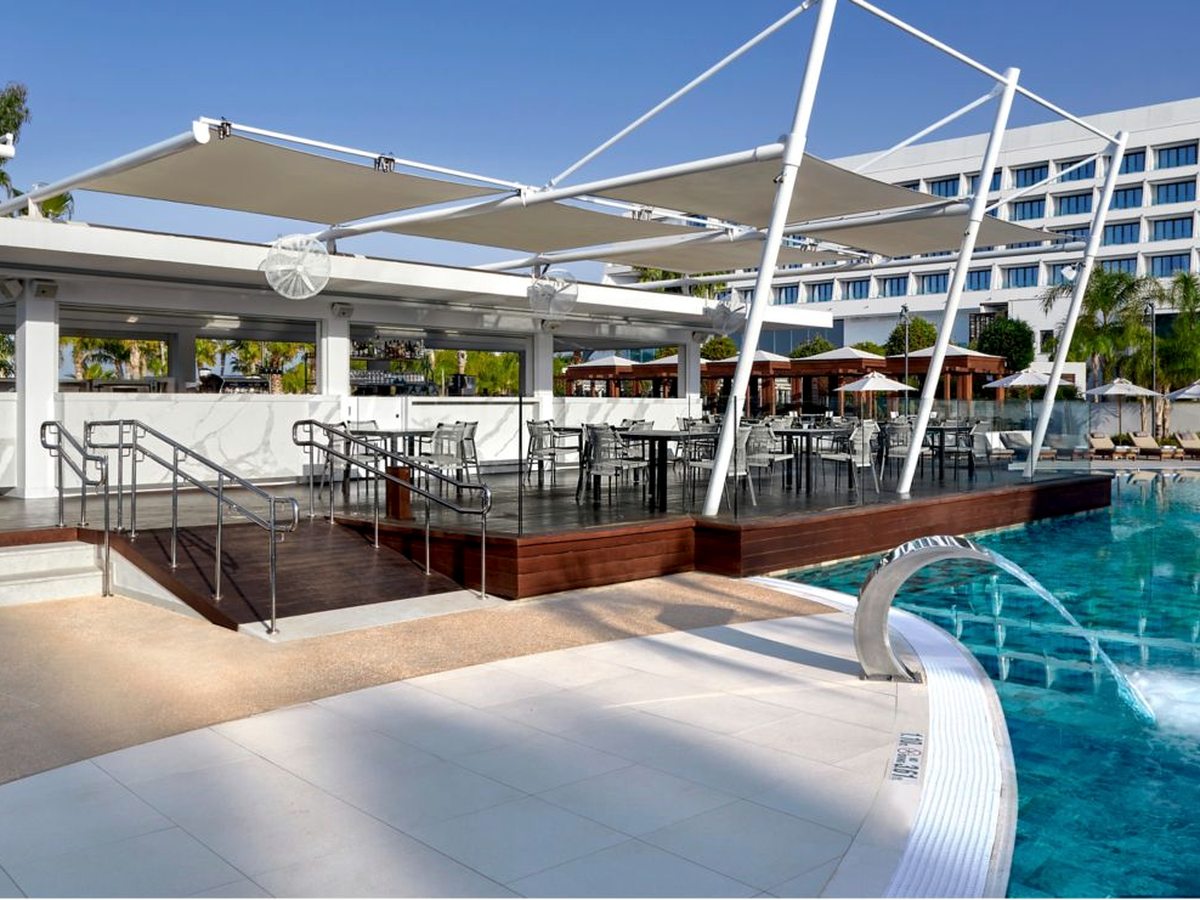 A raised timber deck bar under white sail shades beside a pool, with tables and chairs and the main hotel building behind.