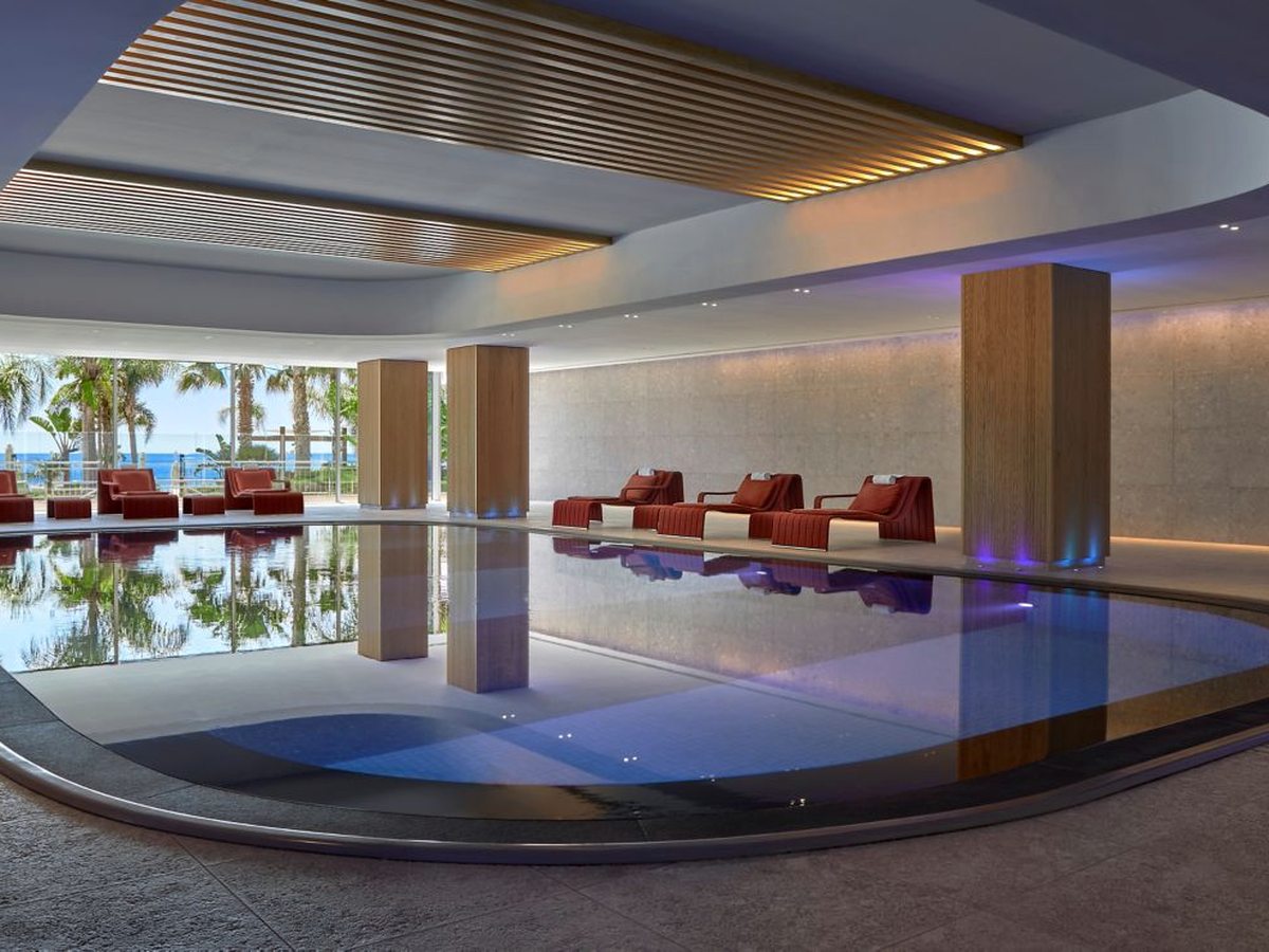 A large curved indoor pool with red loungers along one side, a timber-slatted ceiling and full-height glazing onto palms and the sea.