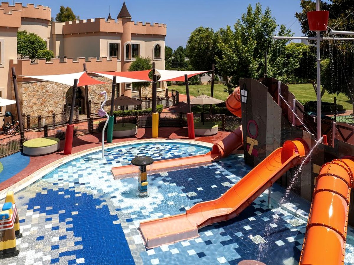 A children's water play area with orange tube slides, tipping buckets and water jets over a shallow tiled pool, with the kids' club castle behind.