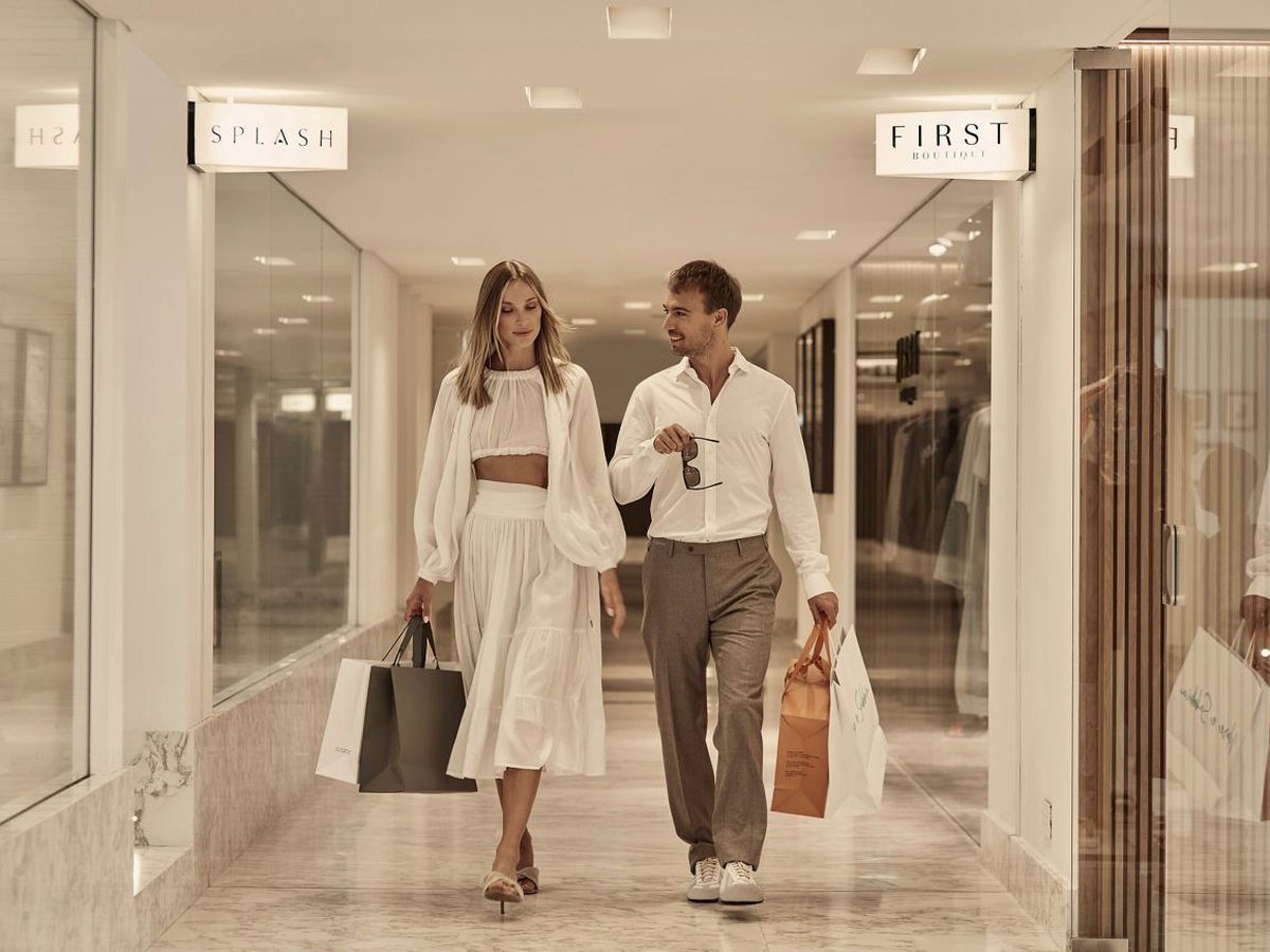 A marble-floored shopping arcade lined with glass-fronted boutiques, with two people walking along it carrying bags.