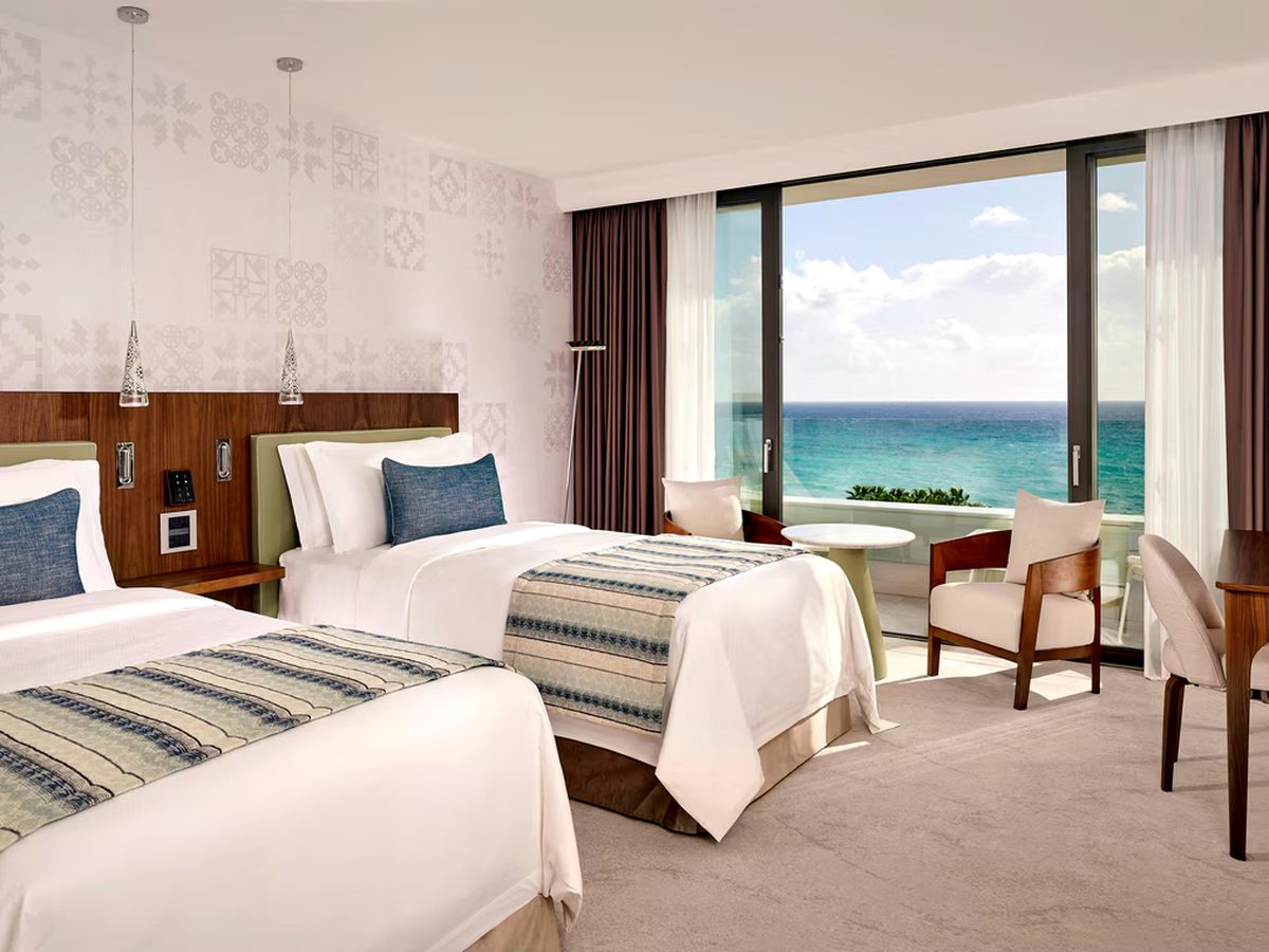 A room with two single beds under a shared wooden headboard, armchairs and a table by sliding doors onto a balcony with an open sea view.