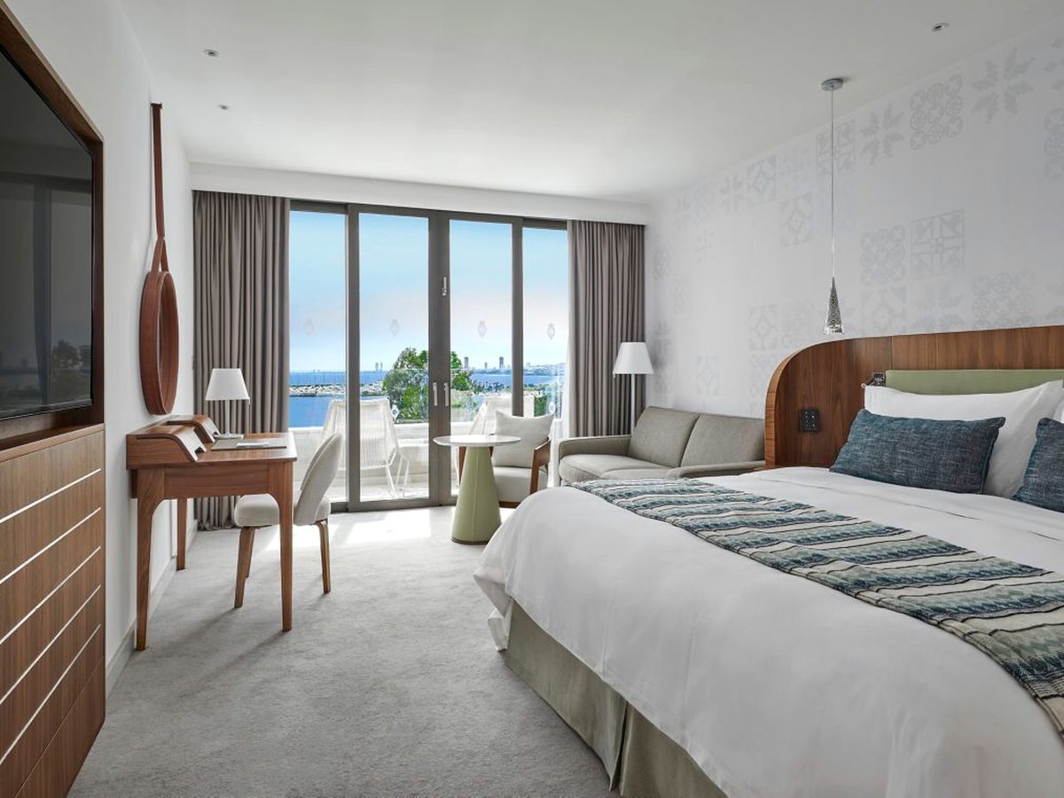 A king bedroom with a curved wooden headboard, a grey sofa, a writing desk and a wall-mounted television, with balcony doors onto a marina view.