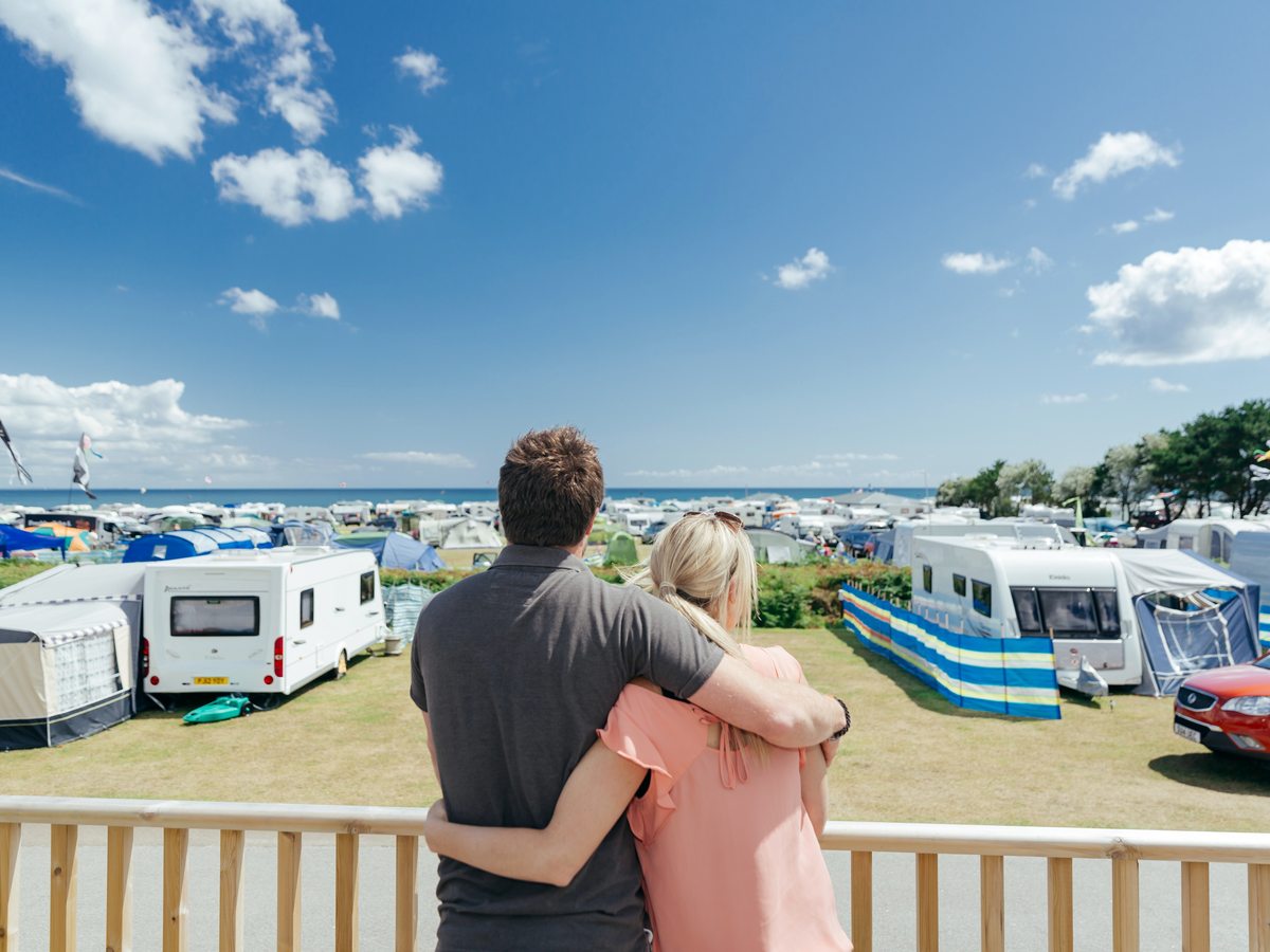 Rows of touring caravans, motorhomes and tents on mown grass under a blue sky with the sea beyond, and a couple standing at a wooden deck rail in the foreground.