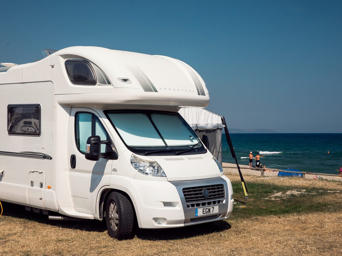 A white motorhome with its awning out, parked on grass at the edge of the beach with the sand and blue sea just beyond.