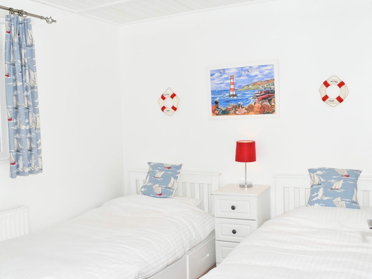 Twin bedroom with two white single beds, blue and white bedding, a bedside table with a red lamp, and framed pictures and lifebelt decorations on the wall.