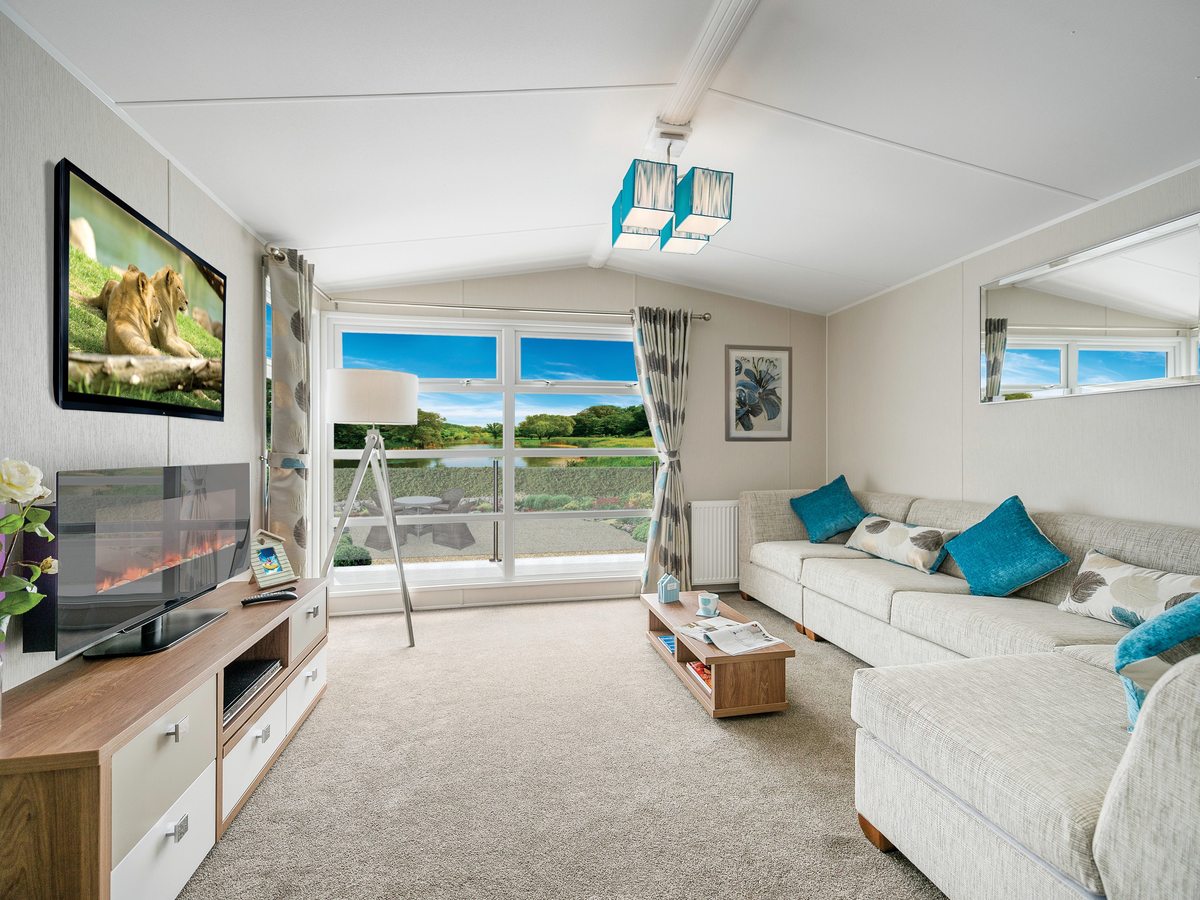 Bright living room with a large cream corner sofa and turquoise cushions, a wall-mounted television, an electric fire and a wide window onto greenery.