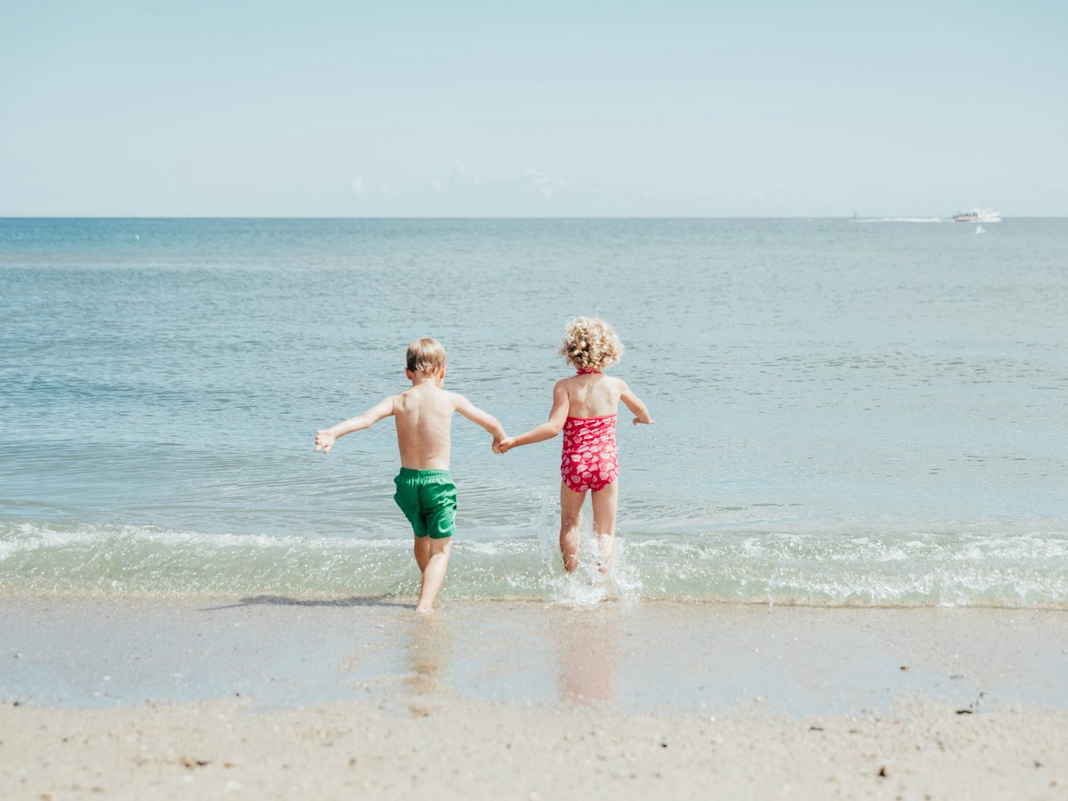Two young children in swimwear holding hands as they run into the shallow water on a sandy beach with a calm sea.