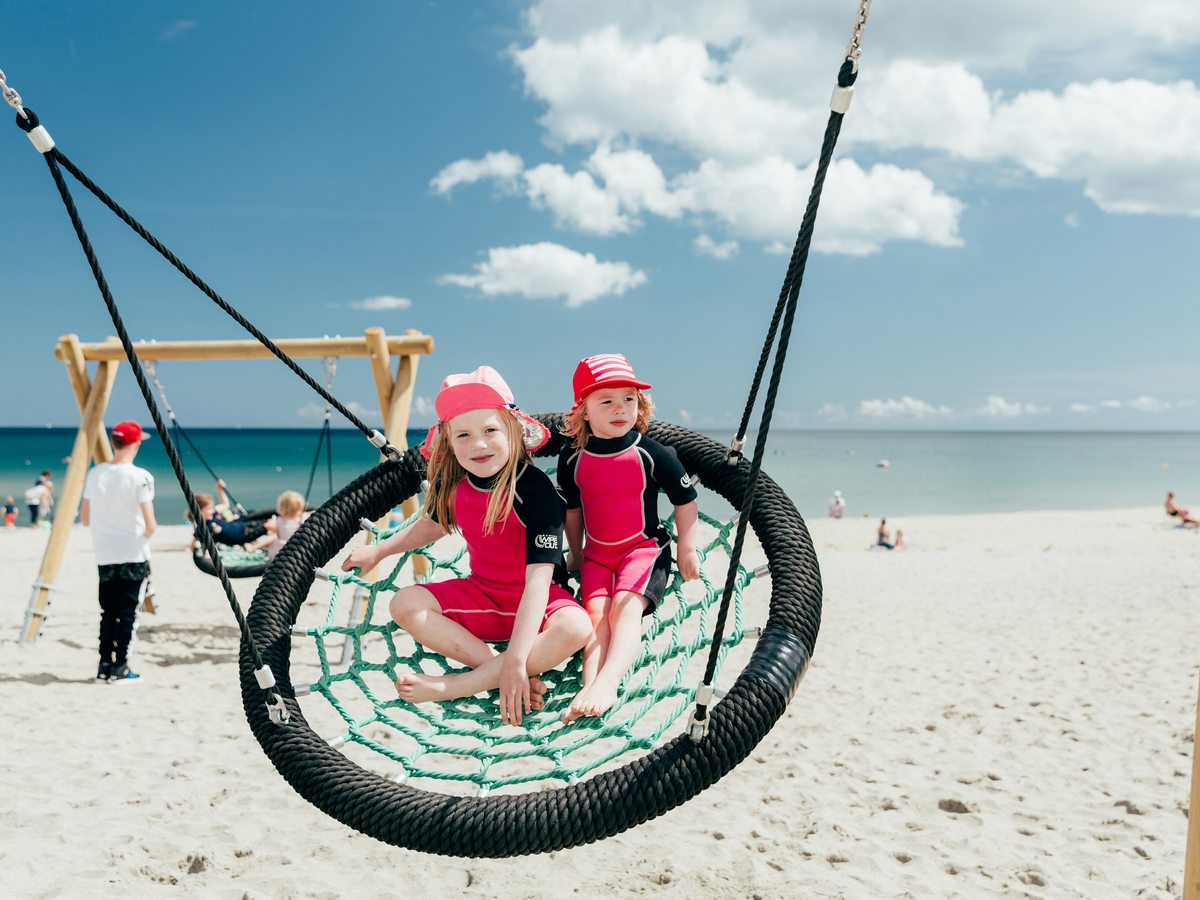 Two small children in sun hats sitting in a large round basket swing on the sand, with the beach and sea behind.
