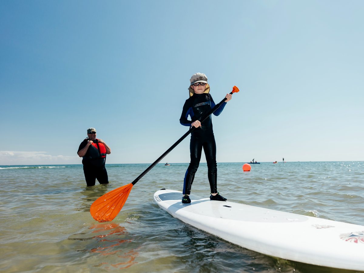 A child in a wetsuit and sun hat standing on a paddleboard in shallow water with an instructor alongside.