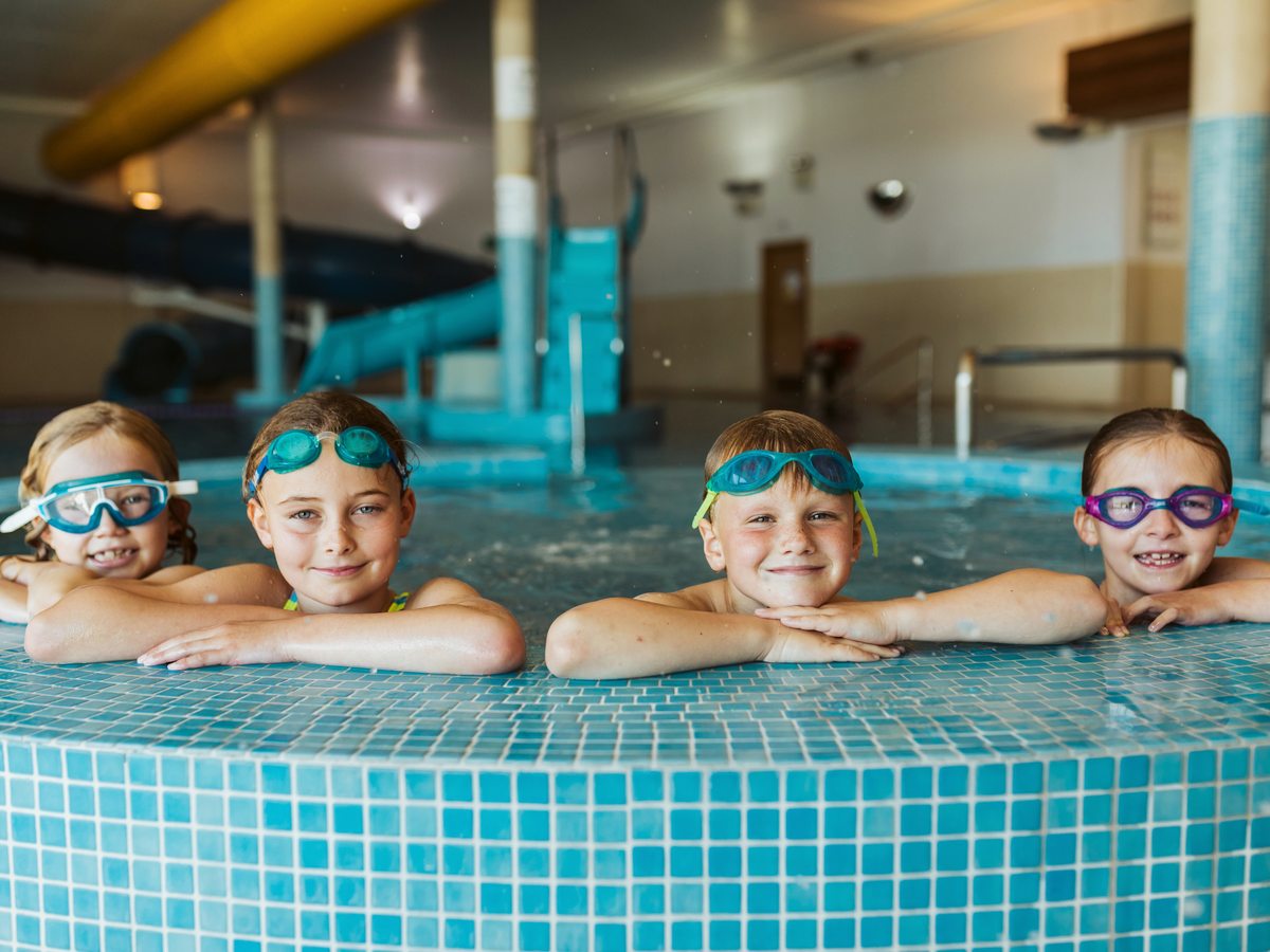 Four children in swimming goggles resting on the tiled edge of an indoor pool, with the flume structure behind them.