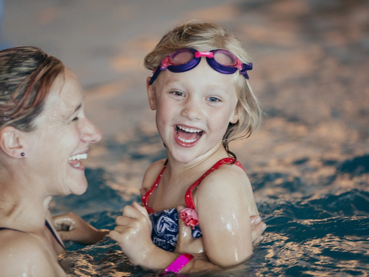 A woman and a young girl in swimming goggles laughing together in the water of the indoor pool.