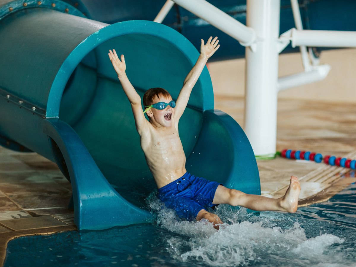 A boy in blue swim shorts coming out of the open end of a teal enclosed water flume into the splashdown channel of the indoor pool.