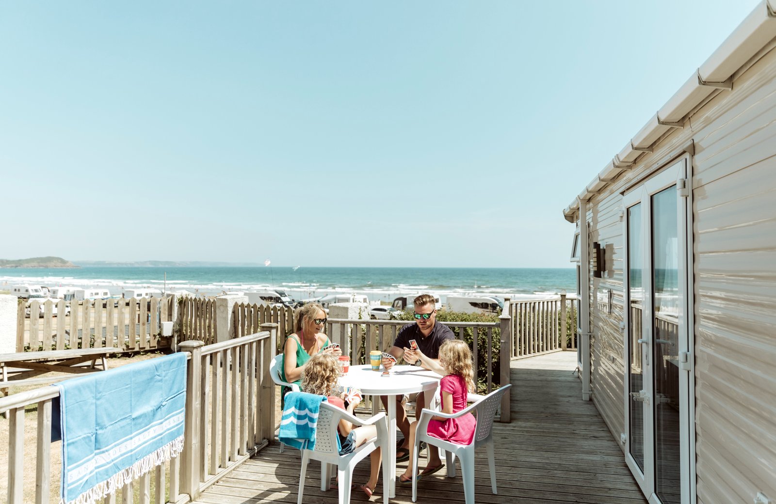A family playing cards at a table on the wooden sundeck of a holiday home, with touring caravans, the sandy beach and the sea behind.