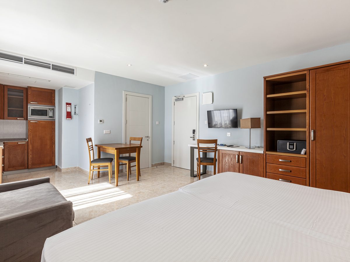 Open-plan studio with a double bed in the foreground, a wooden dining table and two chairs, kitchen units with a microwave along the far wall, and a wardrobe with a safe.