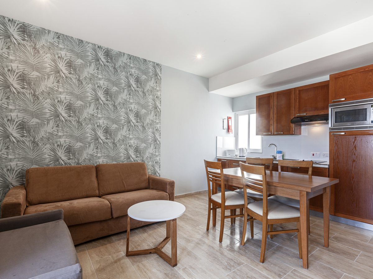 Apartment living room with a brown sofa and a round coffee table against a leaf-patterned wall, and a wooden dining table for four in front of the kitchen units.