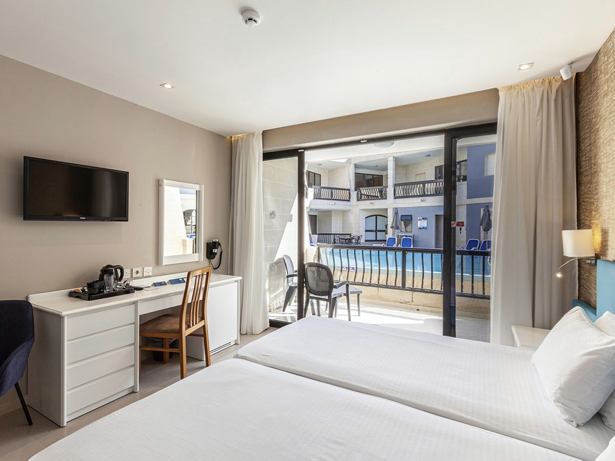 Twin-bedded room with white bedding, a wall-mounted television above a white desk with a kettle tray, and open glass doors onto a balcony above a blue pool courtyard.