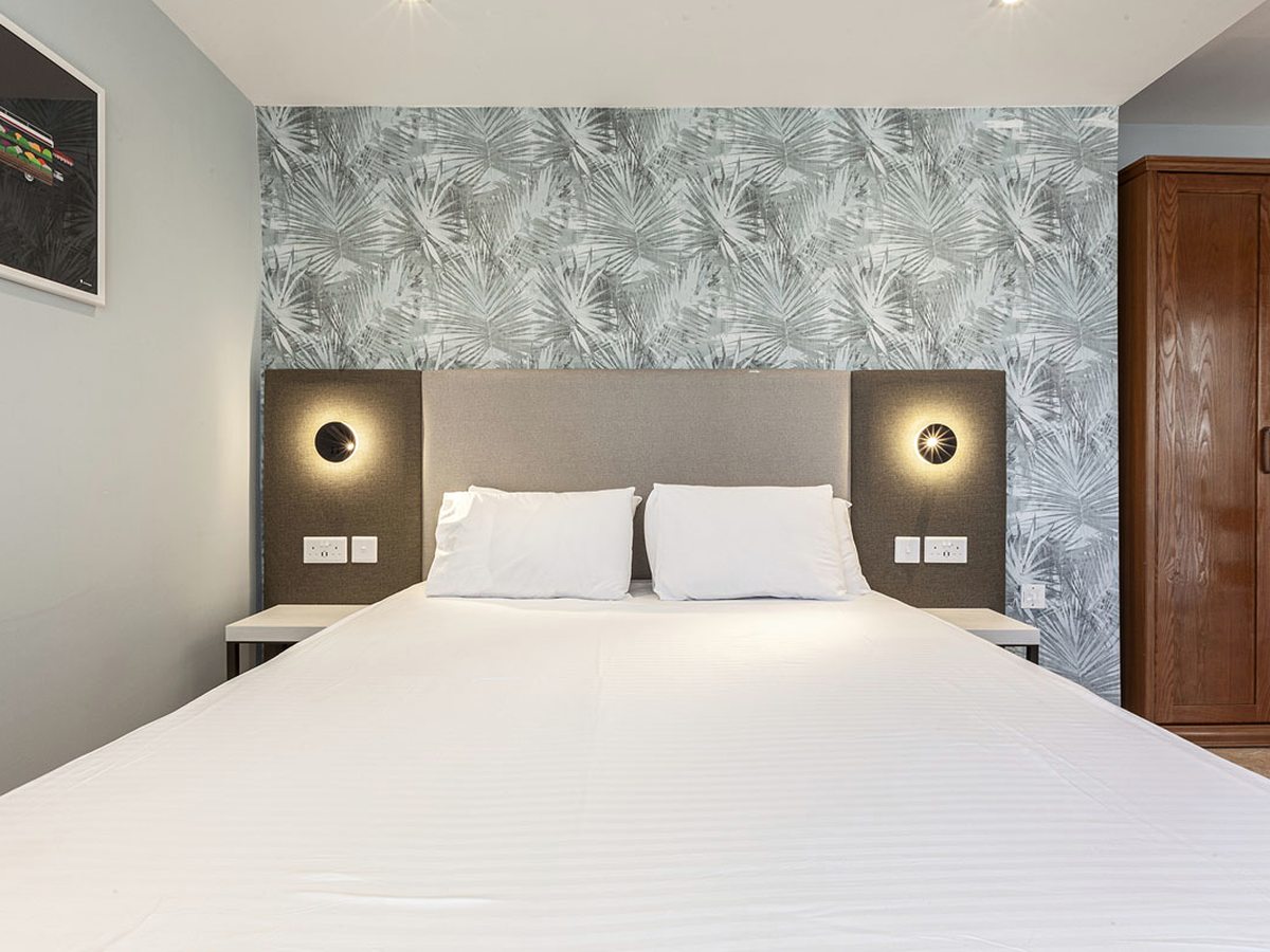 Double bed with a grey padded headboard and round wall lights against a pale leaf-patterned feature wall, with a wooden wardrobe alongside.