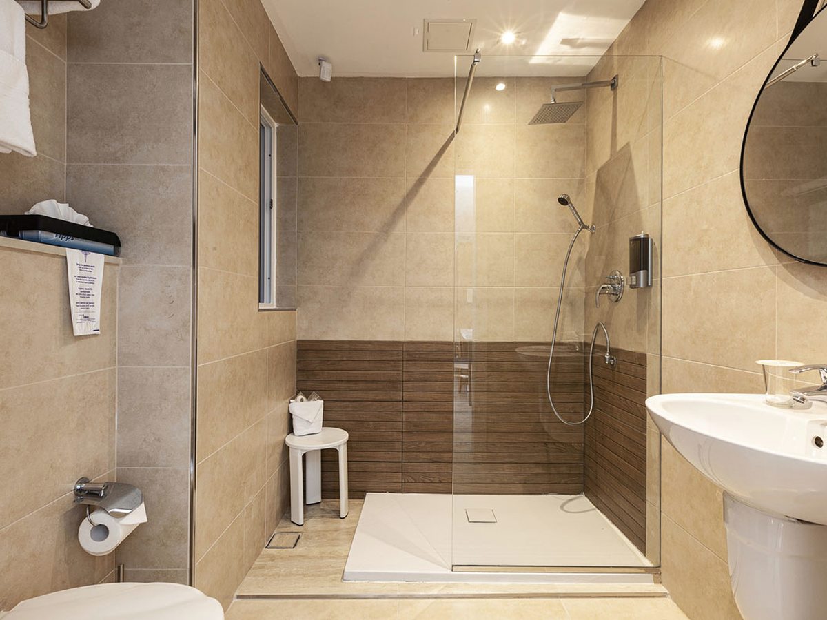 Bathroom with a glass-screened walk-in shower and rain head, a white stool, a basin and a round mirror, on beige and wood-effect tiles.
