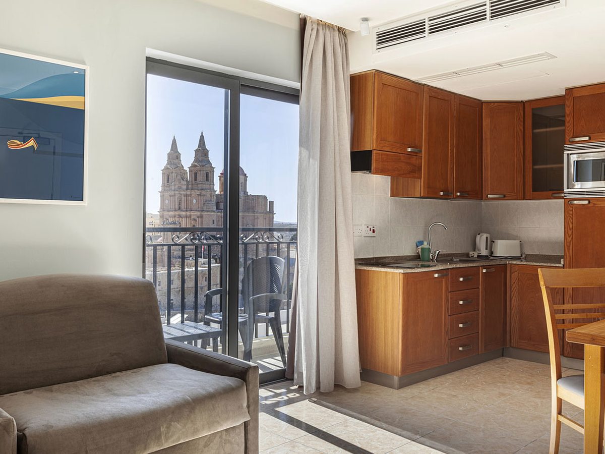 Apartment kitchen with wooden units, a hob, kettle and toaster, beside sliding glass doors onto a small balcony with Mellieħa's twin-towered church in the distance.