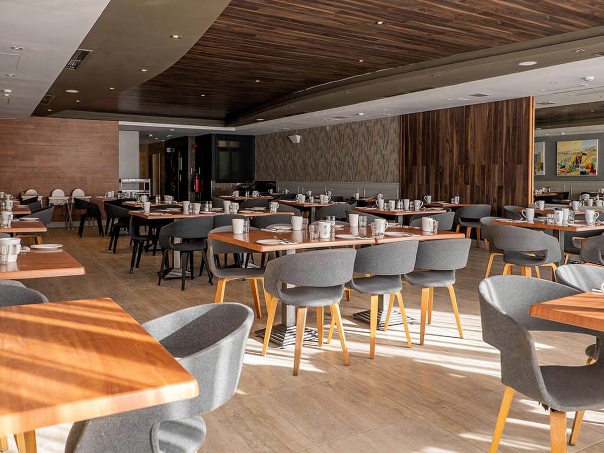 Restaurant dining room with wooden tables, grey tub chairs and a serving counter along the back wall, sunlight falling across the tiled floor.