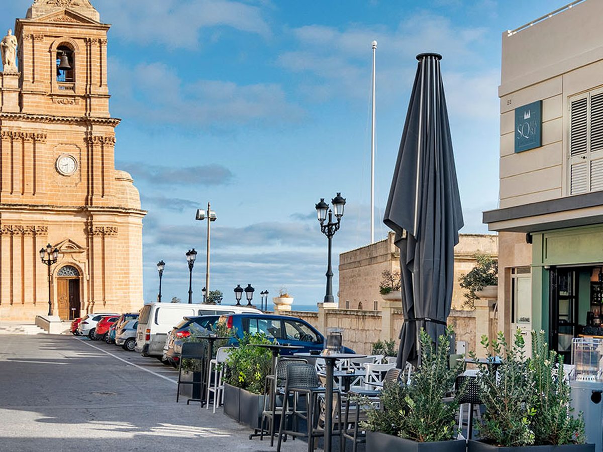 Sunlit square with a honey-stone church and clock tower on the left, parked cars along the middle, and a cafe terrace with planters and a closed parasol on the right, the sea beyond.