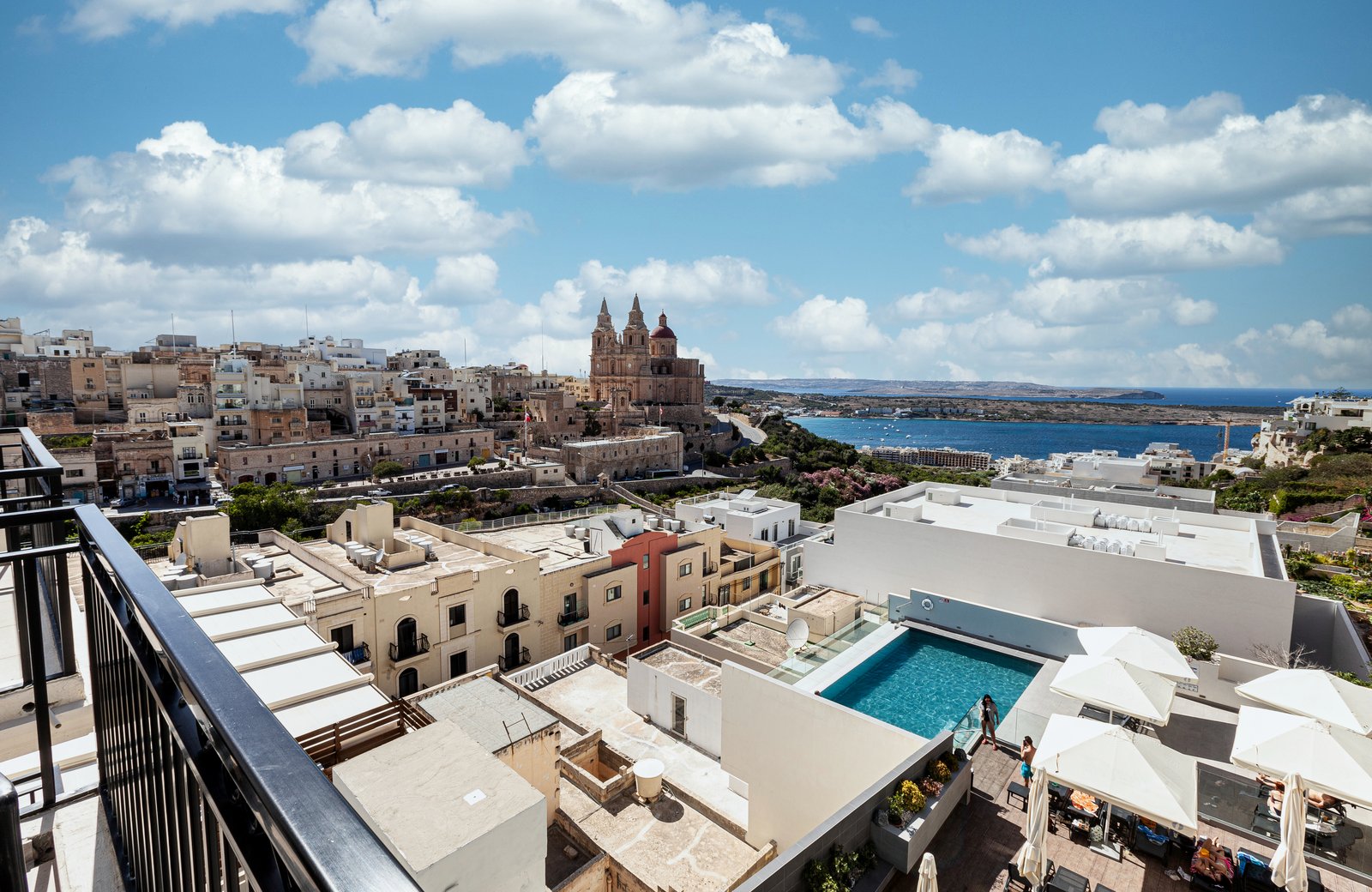 Mellieħa's twin-towered parish church and pale stone houses on the hillside above the bay, with the hotel's pool and white terrace parasols on the rooftops below and the blue sea beyond.