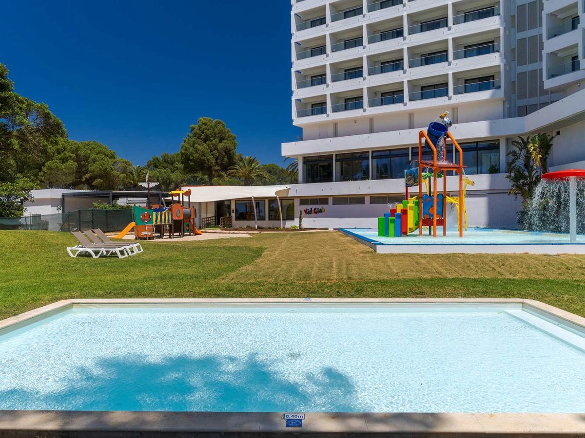 A shallow rectangular children's pool on a lawn, with a colourful water-play tower and a playground next to a Kid Club building and the white resort block behind under a deep blue sky.
