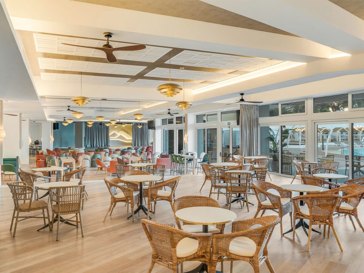 A large lounge of wicker armchairs and round tables under gold pendant lamps and ceiling fans, with a stage at the far end and the pool through the glass doors.