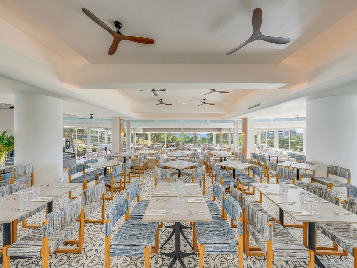 A large bright buffet dining room with rows of set tables, blue woven chairs, a patterned tiled floor, ceiling fans and windows to the gardens and the sea.