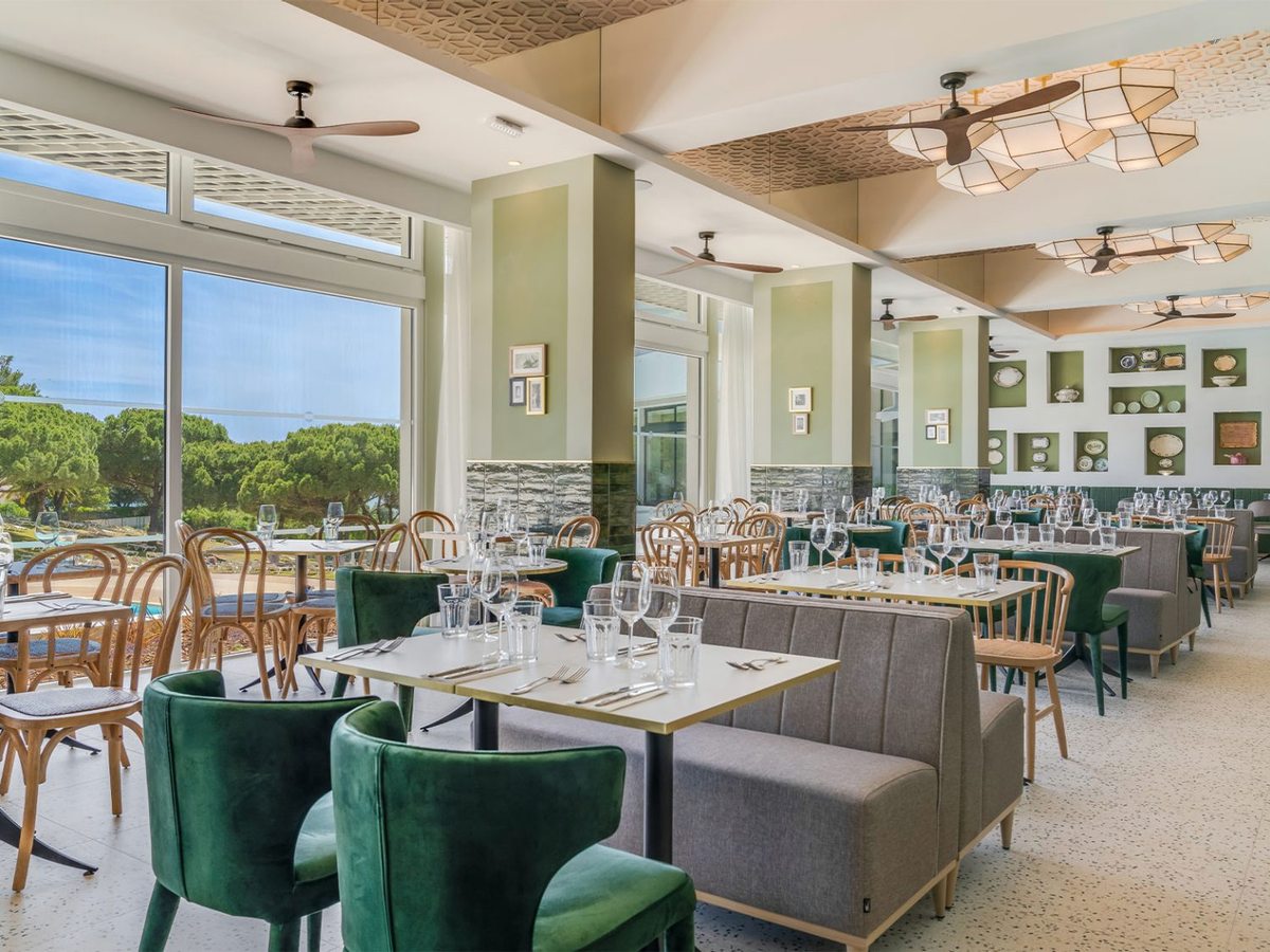 A restaurant with green velvet chairs and grey banquettes, decorative plates on a green wall and full-height windows to an outdoor terrace and pine trees.