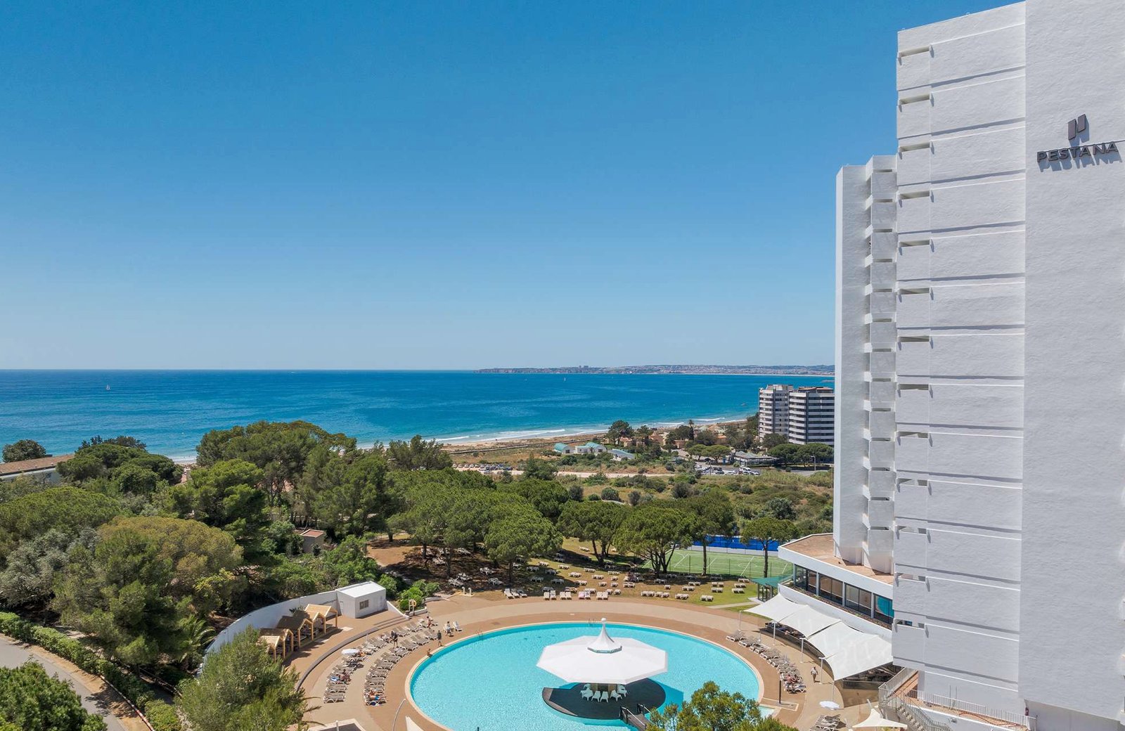 The white Pestana Blue Alvor Beach block above a large circular pool ringed with sun loungers, with pine woods, a sports pitch, the long sandy beach and the blue bay beyond.