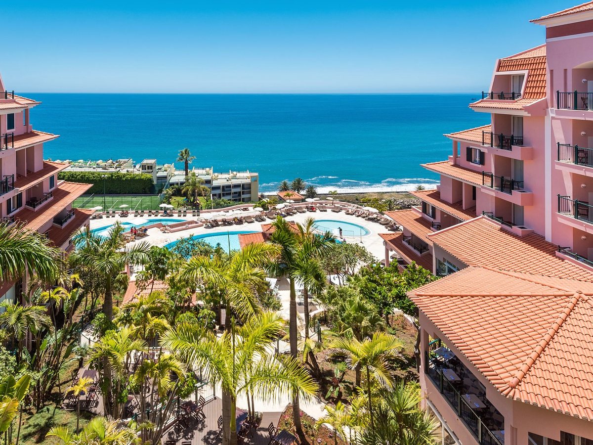 Pink, terracotta-roofed hotel wings around palm gardens, a curved outdoor pool and a smaller round pool on a pale sun terrace, and the blue Atlantic filling the horizon.
