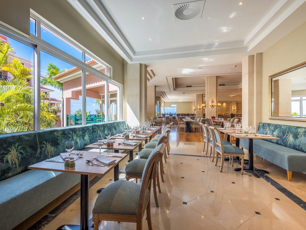 A bright restaurant with green banquette seating, laid tables and wooden chairs on a pale tiled floor, and tall windows onto palms and a pink hotel wing.