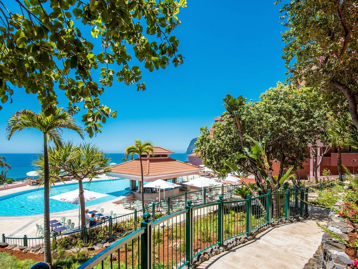 A paved path behind green railings running down through palms and shrubs to a blue pool, a terracotta-roofed poolside pavilion and white parasols, with the Atlantic behind.