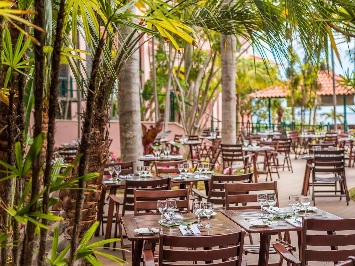 Dark wooden tables and folding chairs laid with glasses on a shaded deck among tall palms, a pink hotel wall on one side and a terracotta-roofed pavilion beyond.