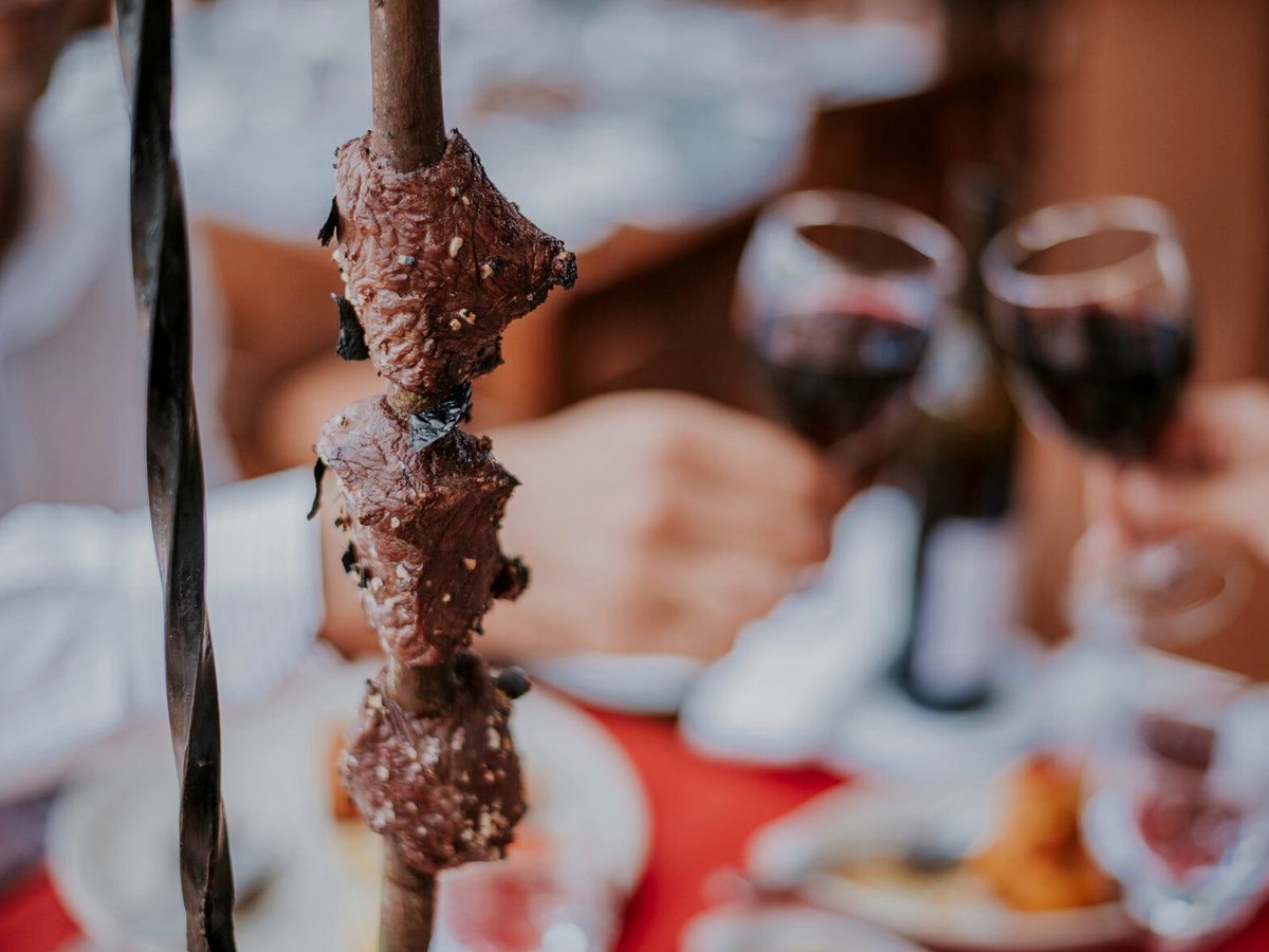 A tall metal skewer of grilled beef standing over a laid restaurant table, with two glasses of red wine and diners out of focus behind.