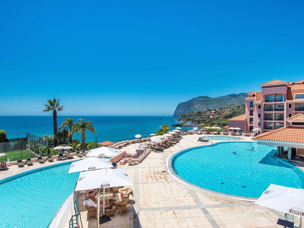 Two curved blue pools either side of a wide stone sun terrace with loungers and white parasols, the pink hotel to the right and the sea and a green headland beyond.