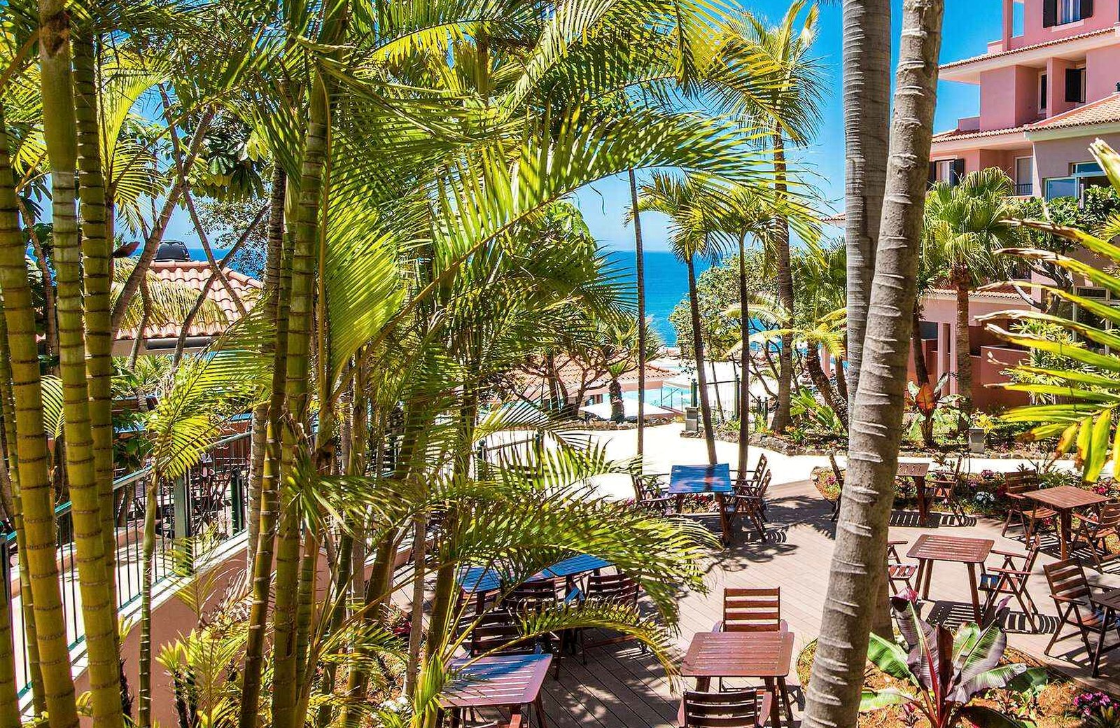 Tall palms and tropical planting around a wooden terrace of tables and chairs in the Pestana Royal's gardens, a pink hotel wing to the right and the Atlantic beyond.