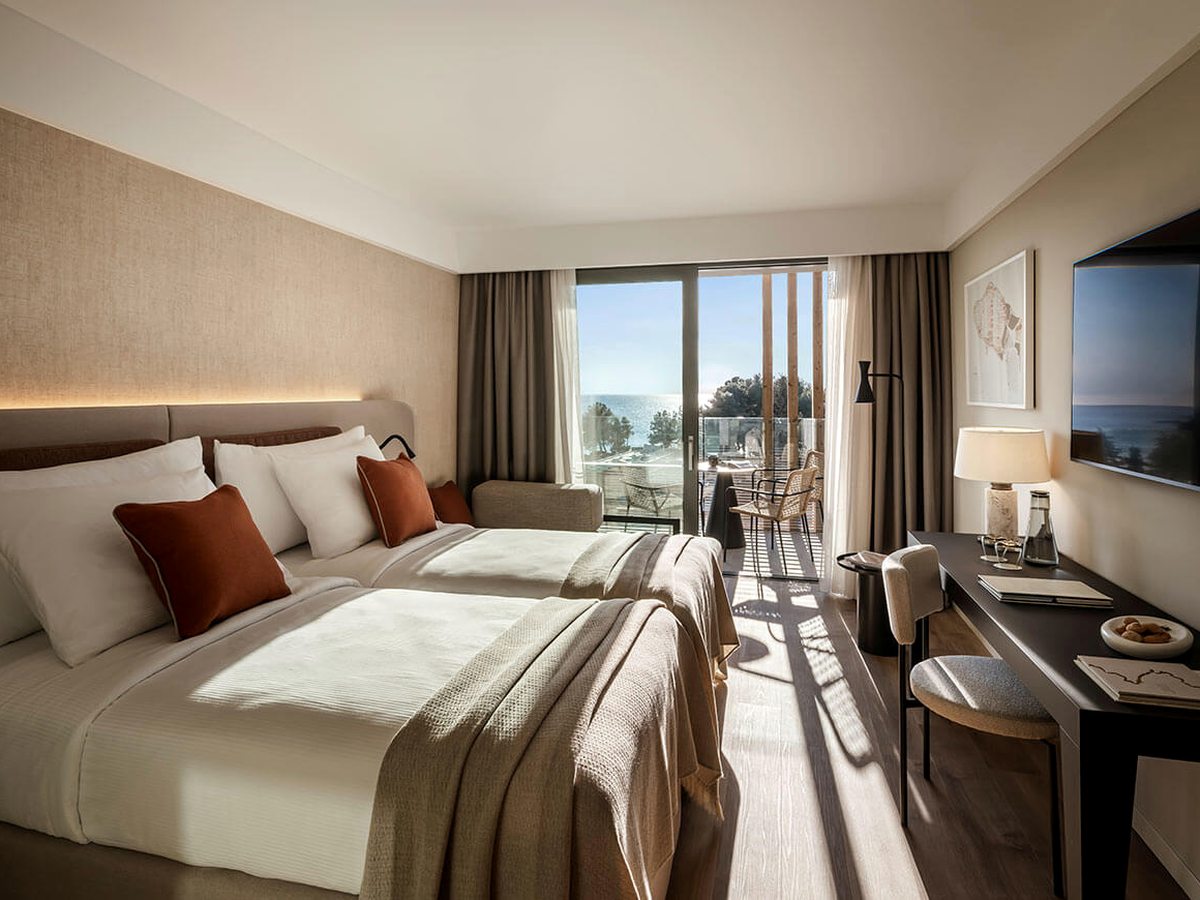 Hotel bedroom with a large bed, rust-coloured cushions, a wall-mounted TV and a desk, and sliding doors onto a balcony with a table and chairs.