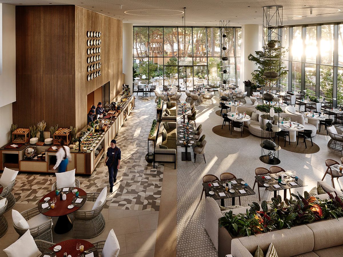 Double-height buffet dining room with wooden serving counters along the left wall, laid tables and armchairs across a patterned floor and a full-height window wall onto the gardens.
