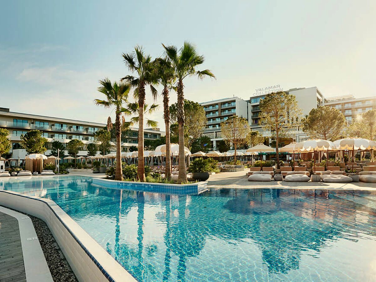 Wide curved outdoor pool with tall palms on a planted island, cream parasols and cabana beds along the far side and the hotel buildings behind.