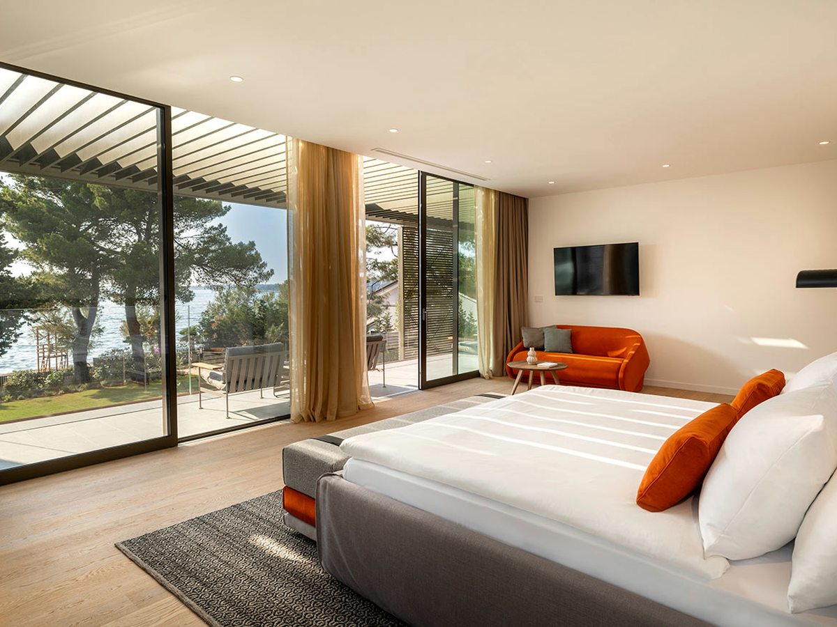 Suite bedroom with a double bed and an orange sofa, and floor-to-ceiling glass onto a private terrace with pines and the sea beyond.