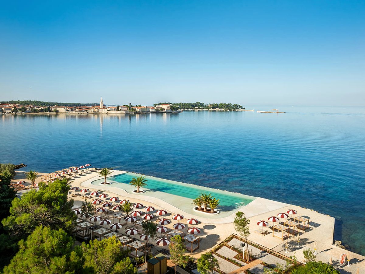 Long rectangular pool set into a paved beach terrace at the water's edge, with rows of red and white striped parasols and loungers, and Poreč old town across the bay.