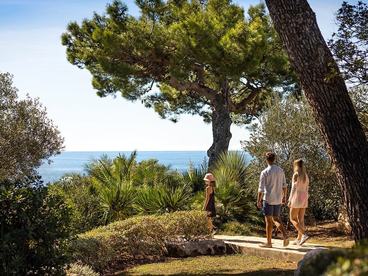 Two adults and a child walking a stone path through pines and olive trees in the resort grounds, with the sea beyond.